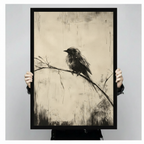 Two hands holding a large black framed vintage charcoal sketch of a lone fluffy black bird perched on a bare diagonal branch with a trailing tail feather and wispy grass marks on a distressed warm cream parchment background monochrome bird oil painting canvas art print, front-facing vertical product size display