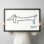 Picasso-inspired minimalist dachshund single-stroke line art in black wood frame held up against a light gray backdrop, white background canvas print