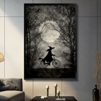 Dark academia witch silhouette riding bicycle through moonlit forest black framed canvas art in moody black and gray on a dark gray paneled wall above a charcoal sofa with pillar candle holders and a black decorative vase