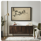 Black framed vintage flying Canada geese botanical illustration canvas art hung above a dark walnut sideboard with stacked books and family photo in a modern living room with gold floor lamp and olive green chair