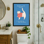 Framed funny whimsical illustration chubby white cat wearing pink ribbed turtleneck sweater with tail raised high and bold handwritten text Welcome To The Bathroom A Cat Will Be With You Shortly on steel blue background canvas print on white wall above white toilet with wicker tray amber candle and trailing potted plant beside marble vanity with brass fixtures and glass shower - humorous bathroom cat sweater welcome wall art for luxury bathroom decor