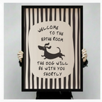 Person holding a large vertical black framed funny bathroom dachshund stripe canvas art print showing bold black and white vertical stripes with a central oval featuring hand-lettered welcome to the bathroom above a jumping cartoon dachshund silhouette and the humorous quote the dog will be with you shortly against a light gray background