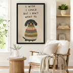Black framed funny dachshund I Wish I Could But I Don't Want To canvas art with a grumpy black dachshund wearing a colorful striped knit sweater on a warm beige background, displayed beside a white linen armchair in a cozy reading nook
