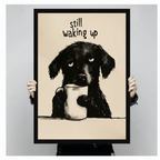 Person holding a black framed humorous pencil illustration drowsy black dog clutching white coffee mug "Still Waking Up" art print on warm beige background against a light gray backdrop