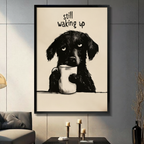 Funny illustration sleepy black dog holding white coffee mug with drowsy eyes "Still Waking Up" quote black framed canvas art on warm beige background on a dark gray paneled wall above a charcoal sofa with brass candle holders and a black decorative vase