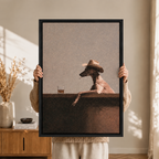 Person holding a black-framed humorous canvas print featuring a greyhound dog in a cowboy hat beside a whiskey glass, warm bohemian interior background