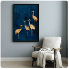 Black framed gold foil crane quartet canvas art print with four graceful golden cranes on a deep navy blue textured background on a light blue-grey wall beside a white linen armchair with draped throw