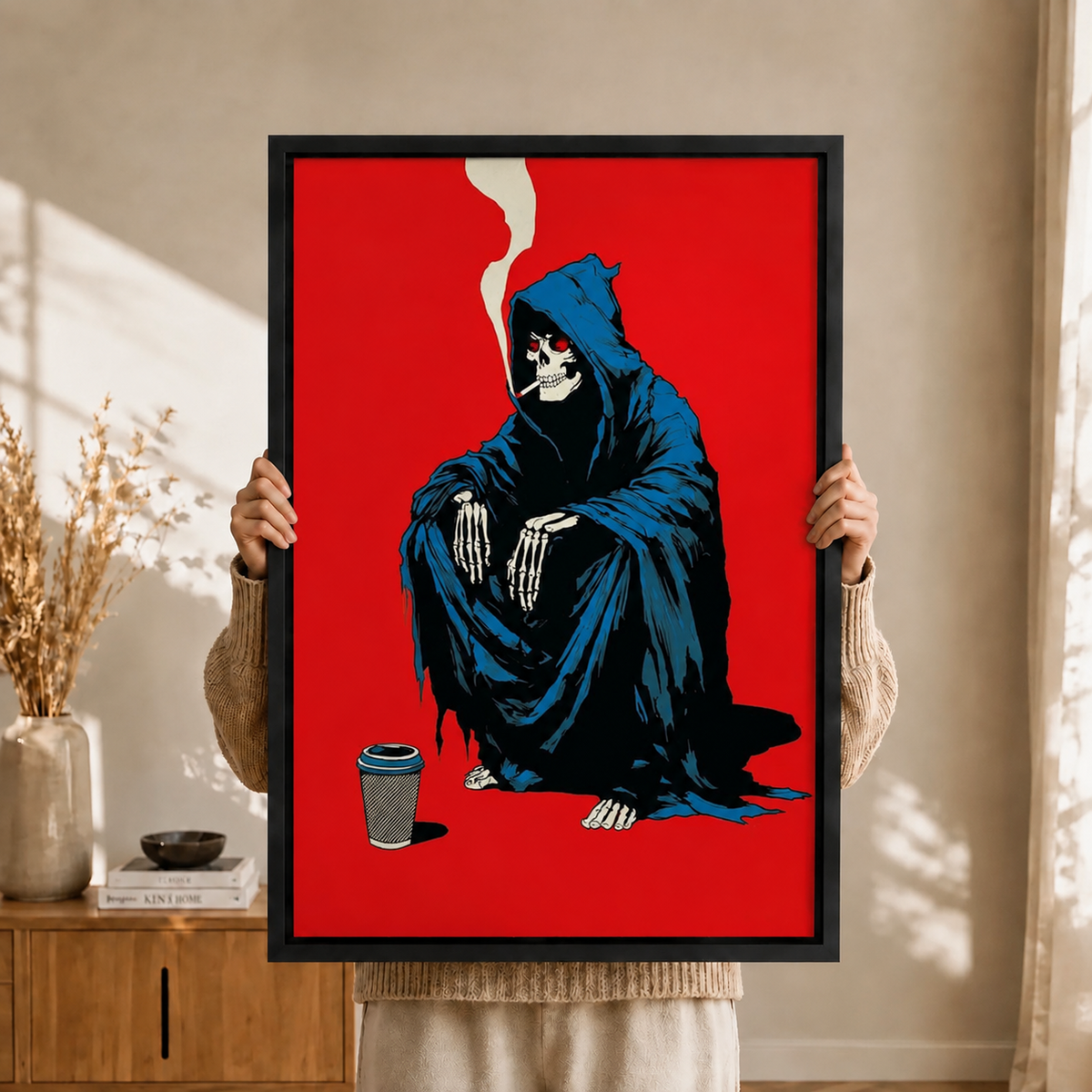 Person holding a black-framed graphic wall art canvas featuring a dark humor grim reaper skeleton in a blue robe smoking and sitting beside a coffee cup against a striking red background, bohemian interior setting
