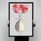 	Hand-held product shot of a black-framed art print showing a cute hedgehog with rosy cheeks clutching a bouquet of red and pink heart balloons