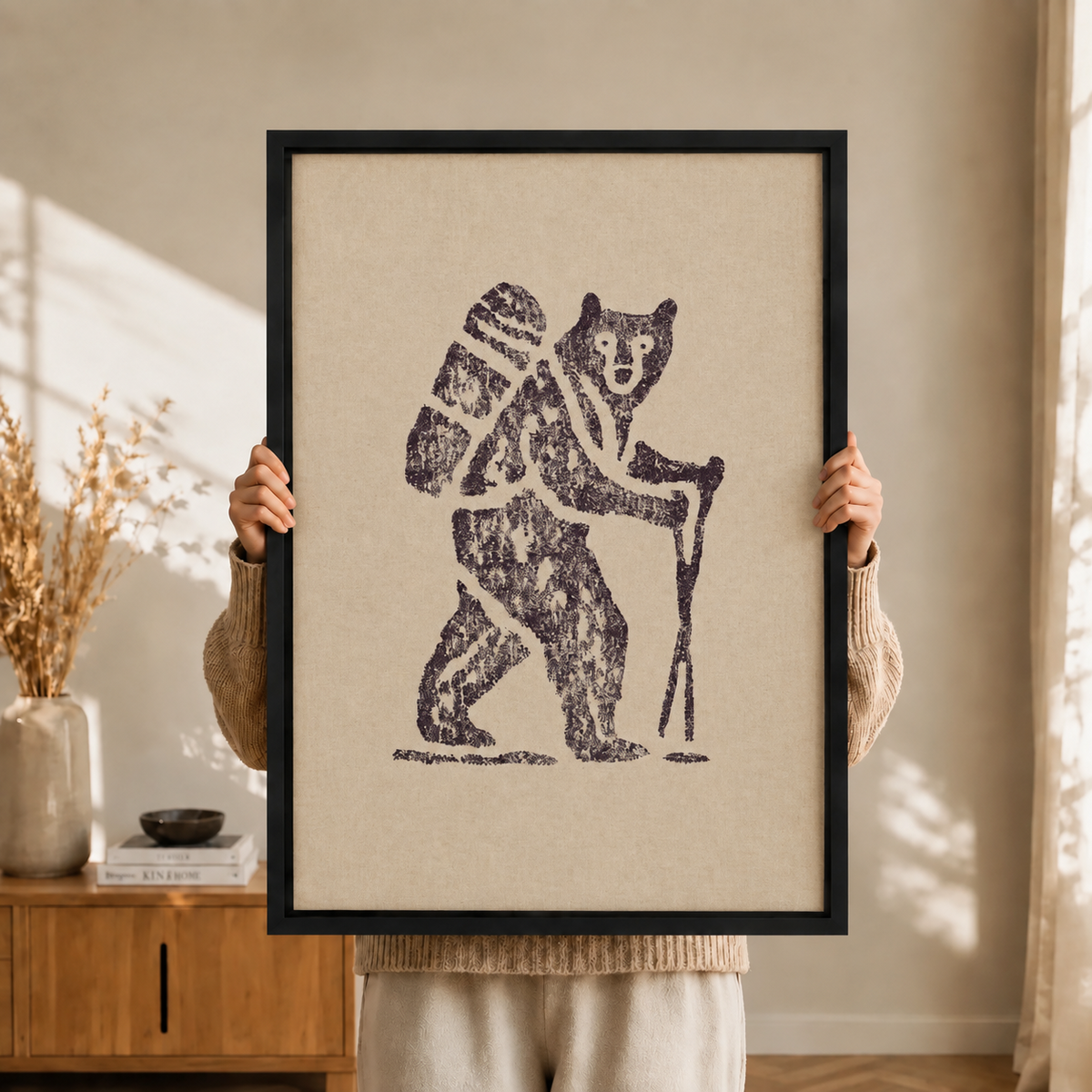 Person holding a black-framed canvas art print of a bold linocut bear carrying a hiking backpack and walking stick, warm bohemian interior background