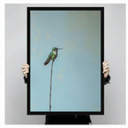Two hands in black jacket sleeves holding a large black framed minimalist green hummingbird perched on a tall thin branch canvas art print with soft blue background, front-facing vertical product size display