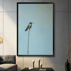 Black framed minimalist hummingbird perched on a tall thin branch canvas wall art with soft powder blue textured background displayed in a modern dark gray living room with charcoal sofa and gold candlesticks