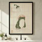 Black framed Japanese woodblock frog shower canvas art with a plump dark green frog wearing a pink ruffled shower cap standing under a vintage rainfall showerhead in a tiled shower with a red seal stamp in the lower left on a warm aged parchment background, displayed above a minimalist bathroom sink with a black faucet, soap dispenser, and small potted plant