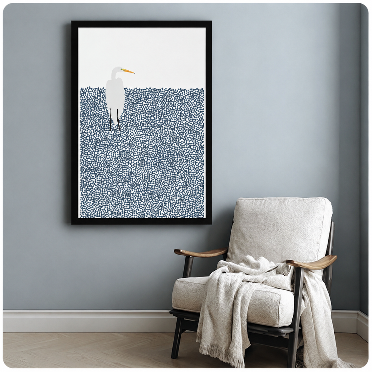 Black framed minimalist white egret with yellow beak standing in dotted navy blue water art print on a blue-grey wall beside a white linen armchair with draped throw