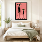 Black framed retro atomic-age black cat and green frog in suits canvas art with a tall slender black cat in a black suit and white tie beside a short green frog in matching attire on a coral pink background, hung above a wooden bed frame in a sun-drenched Scandinavian bedroom with crisp white linen bedding,