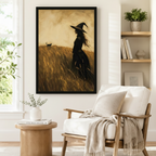 Black framed vintage witch and cat wheat field canvas art showing a long-haired witch in a pointed hat standing in a sunlit golden field with a black cat strolling through tall grass in warm sepia oil tones, displayed on a white wall beside a light upholstered wooden armchair draped with a cream fringed throw blanket near a shelf of plants
