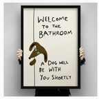 Person holding large black framed funny dog bathroom wall art print with welcome to the bathroom a dog will be with you shortly quote